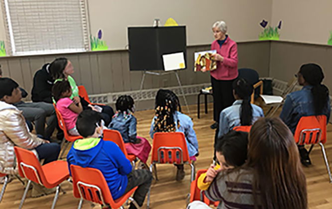 A woman holding visual aid teaches a group of students seated in chairs.