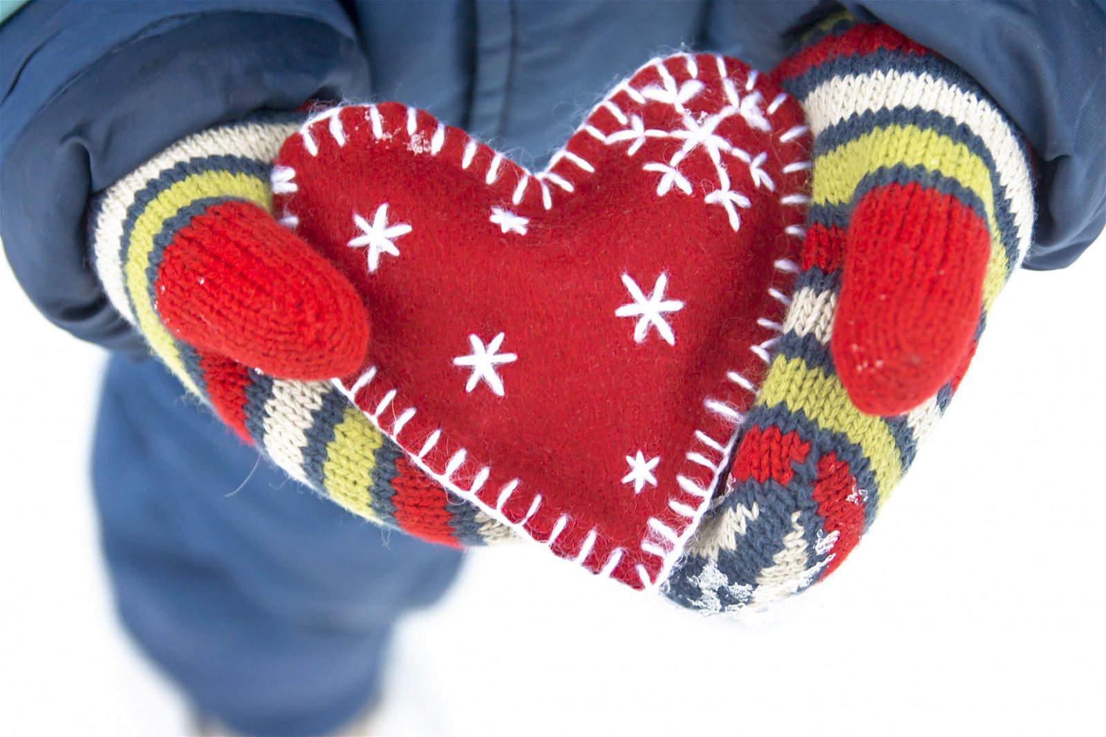 A child's mittened hands are holding a red felt heart embroidered with snow flakes.