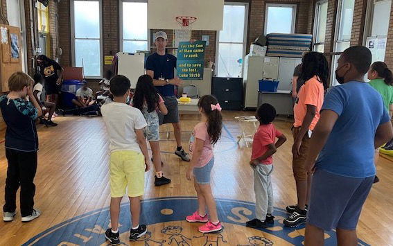 A man stands in front of a group of standing children. He's holding a verse visual and leading them in saying the verse