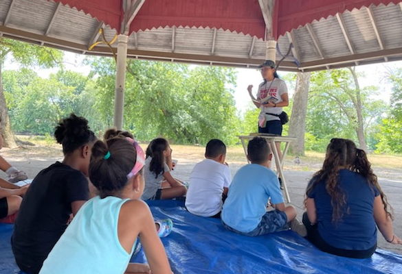 A teacher stands in front of a group of attentive children seated on a tarp.