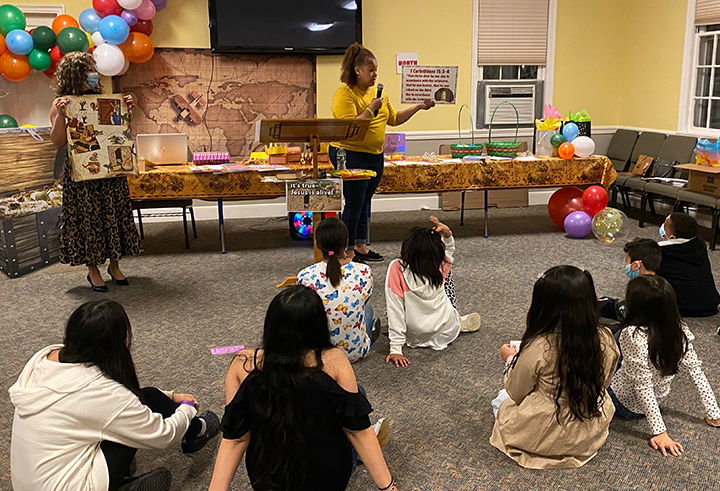 A woman teaches a group of children seated on a carpeted floor. There are balloons and treasure chests in the background.