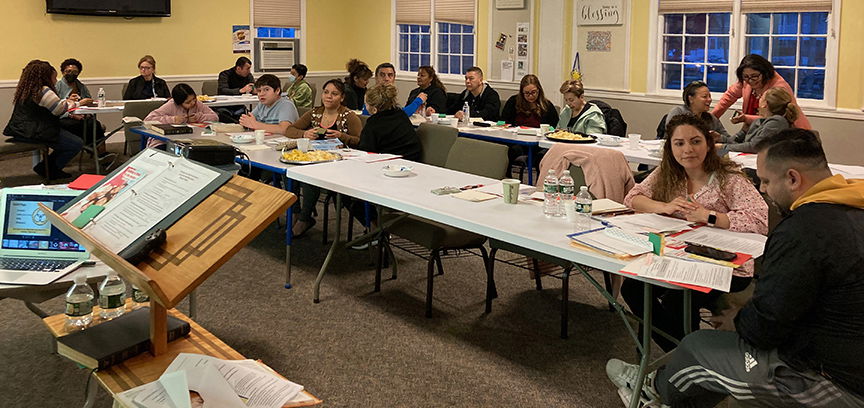 Teachers seated at long tables practice presenting to each other. The instructors notes are visible at lower left.