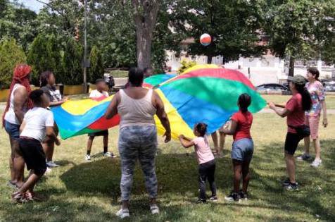 Children and adults play, tossing a ball in the air, with a colorful parachute.